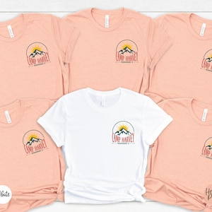 K&ouml;nnte beinhalten: F&uuml;nf kurz&auml;rmelige T-Shirts mit Rundhalsausschnitt in einer Gruppe angeordnet. Vier T-Shirts sind pfirsichfarben meliert und eines ist wei&szlig;. Jedes T-Shirt hat eine kreisf&ouml;rmige Grafik auf der linken Brust mit einem Gebirge, einer aufgehenden Sonne und dem Text Camp Harvey Fredericksburg, TX.