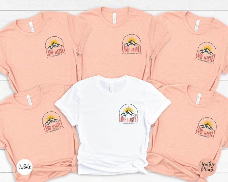 K&ouml;nnte beinhalten: F&uuml;nf kurz&auml;rmelige T-Shirts mit Rundhalsausschnitt in einer Gruppe angeordnet. Vier T-Shirts sind pfirsichfarben meliert und eines ist wei&szlig;. Jedes T-Shirt hat eine kreisf&ouml;rmige Grafik auf der linken Brust mit einem Gebirge, einer aufgehenden Sonne und dem Text Camp Harvey Fredericksburg, TX.