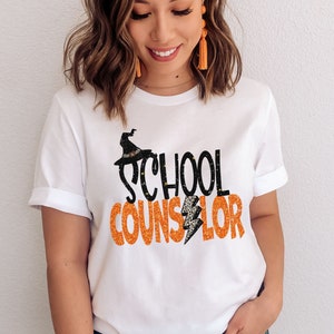 May include: White t-shirt with the text "SCHOOL COUNSELOR" in orange glitter letters with a black witch hat and lightning bolt graphic.