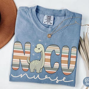 May include: A light blue t-shirt with a striped design that spells out "NCU nurse" with a cartoon dinosaur in the middle of the letters.