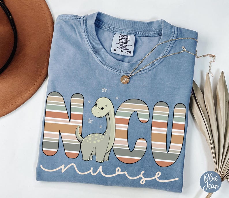 May include: A light blue t-shirt with a striped design that spells out "NCU nurse" with a cartoon dinosaur in the middle of the letters.