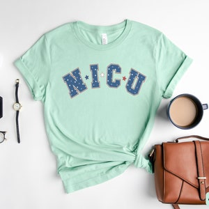 May include: A light green t-shirt with the text "NICU" in blue and red lettering with white stars. The letters are outlined in white.