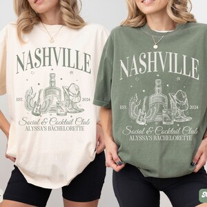 K&ouml;nnte beinhalten: Zwei T-Shirts mit einem grafischen Design, das den Text "Nashville" und eine Zeichnung einer Flasche, einer Gitarre und Cowboystiefeln zeigt. Der Text "Social & Cocktail Club" und "Alyssa's Bachelorette" sind ebenfalls auf den Shirts enthalten. Die Shirts sind in Elfenbein und Moosgr&uuml;n.