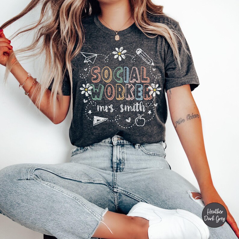 May include: Dark gray t-shirt with the words "SOCIAL WORKER mrs. smith" in colorful letters. The design includes a paper airplane, flowers, a pencil, a ruler, and an apple. The shirt is made of a soft material.