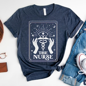 May include: A navy blue t-shirt featuring a white graphic of a tarot card design. The card depicts a caduceus symbol with the text "The Nurse".
