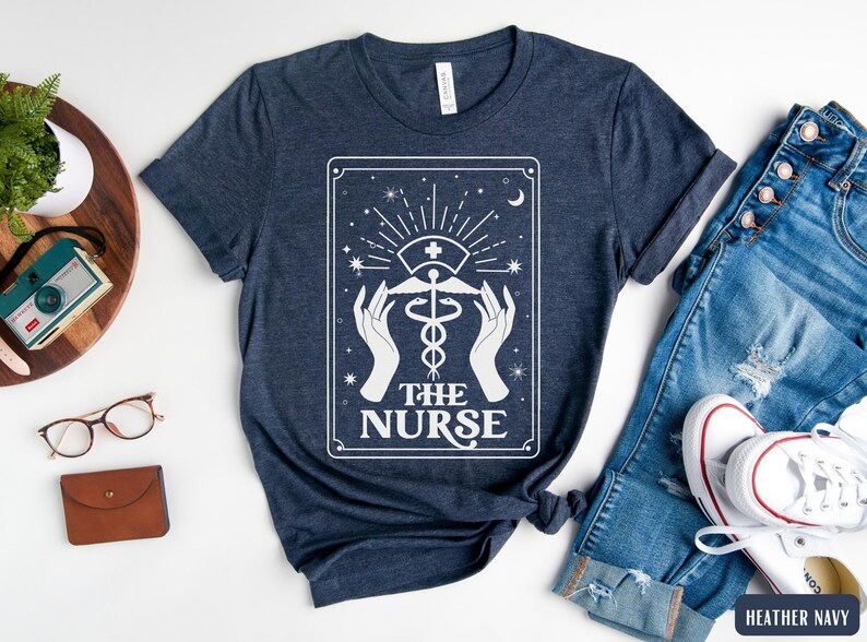 May include: A navy blue t-shirt featuring a white graphic of a tarot card design. The card depicts a caduceus symbol with the text "The Nurse".