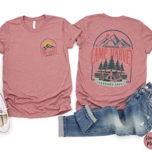 K&ouml;nnte beinhalten: Ein kurz&auml;rmeliges T-Shirt in Mauve-Melange mit einer kleinen Grafik auf der Vorderseite und einer gro&szlig;en Grafik auf der R&uuml;ckseite. Die Motive zeigen Berge, Tannen und ein Zelt mit dem Text Camp Harvey, Fredericksburg, TX, und January 2024 in Rot und Gr&uuml;n.