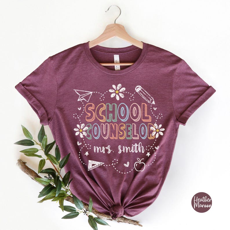 May include: Heather maroon t-shirt with the words "SCHOOL COUNSELOR" in colorful letters, along with a pencil, paper airplane, flowers, apple, and the name "mrs. smith".