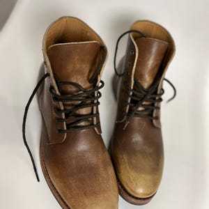 May include: A pair of brown leather boots with black laces. The boots have a worn, distressed appearance, with a lighter shade of brown on the toes. The soles are a darker brown with a red edge. The boots are sitting on a white surface.