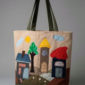 Può includere: Borsa tote beige con una scena di villaggio applicata stravagante. Il design include case con tetti blu, rossi e gialli, un albero, un sole e nuvole. La borsa ha manici verdi e una base verde.