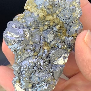 May include: A close-up of a mineral specimen featuring a cluster of metallic, silver-gray galena crystals. The galena is interspersed with small, tan-colored crystals. The specimen is held in a hand against a dark background.