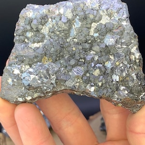 May include: A hand holds a piece of raw, metallic-looking mineral. The surface is a mosaic of small, reflective, gray and silver crystals. The overall shape is irregular, with a rough, textured appearance. The background is dark.