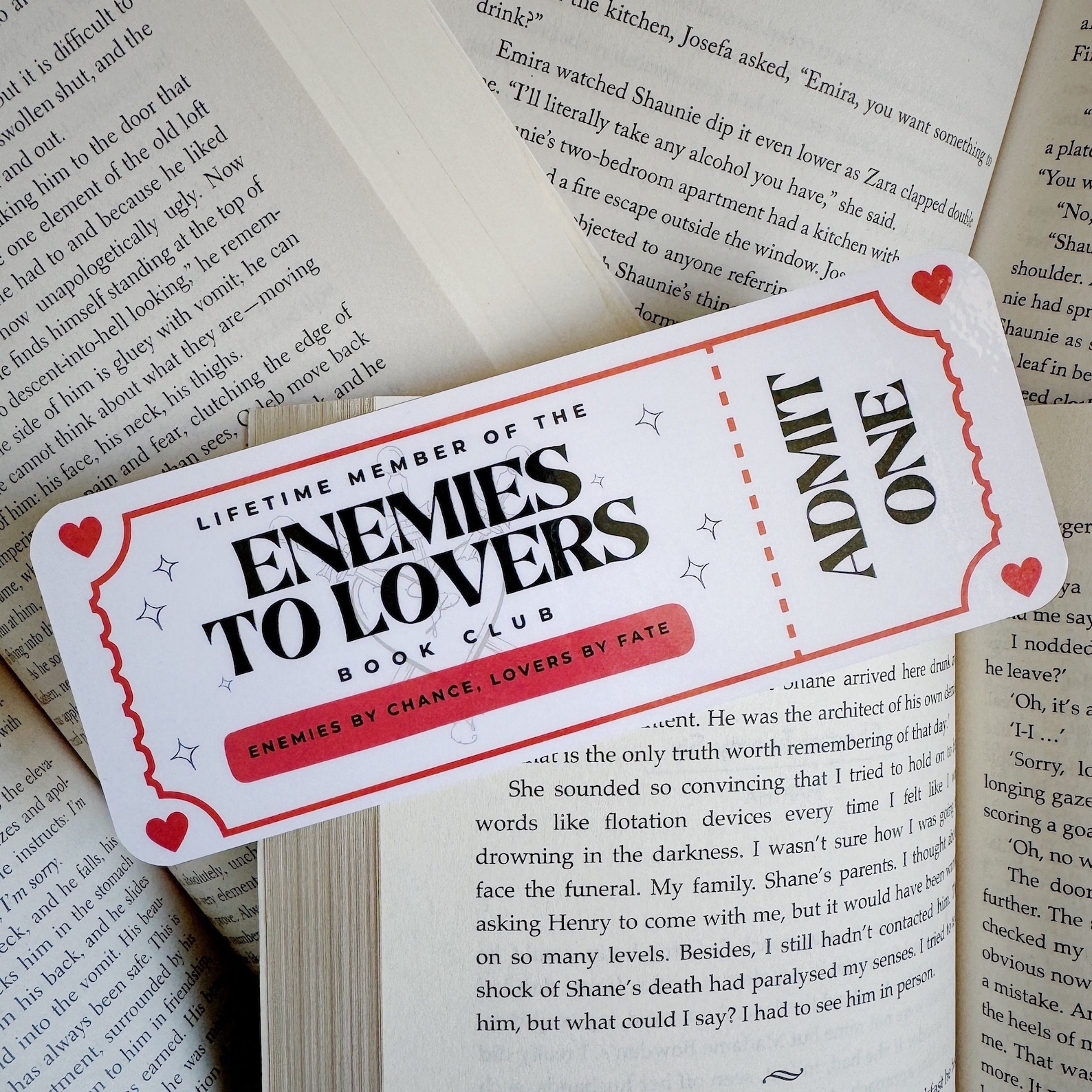 Several books open with a bookmark on top shaped like a movie ticket that says "lifetime member of the enemies to lovers book club."