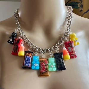 May include: A silver chain necklace with various candy charms, including miniature candy bars, gummy bears, and candy corn. The charms are in vibrant colors, such as red, blue, and yellow.