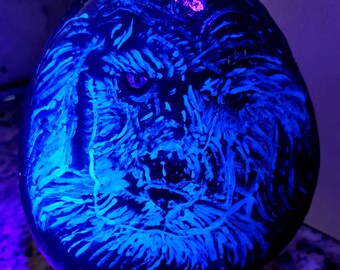May include: A black stone painted with a white wolf face, glowing blue under a black light. The stone is on a black metal stand.