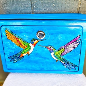 May include: A bright blue mailbox with a hinged lid, featuring two colorful hummingbird illustrations. The hummingbirds have vibrant wings in shades of orange, yellow, green, and purple, set against a light blue background. A silver lock is centered on the front.