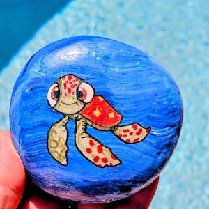 May include: A hand-painted, round stone featuring a cartoon sea turtle design. The turtle has a red shell with yellow spots, green flippers, and a smiling face. The stone is painted with blue water-like patterns.