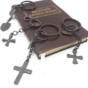 May include: Three dark metal keychains featuring cross pendants and small shield-shaped charms, resting on a brown book titled BELOVED CATHOLIC PRAYERS. Each keychain includes a circular ring and a short chain connecting to the religious symbols.