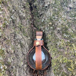 Puede incluir: Una botella de vidrio redonda con un tap&oacute;n de corcho est&aacute; envuelta en cuero marr&oacute;n con una hebilla de lat&oacute;n. La botella est&aacute; colgada de un tronco de &aacute;rbol.