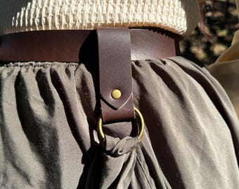 May include: A brown leather belt with a metal ring and a brown leather strap. The belt is worn around the waist and the strap is attached to the ring.