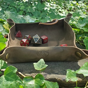 Puede incluir: Un dado de cuero marrón con un diseño decorativo en la parte inferior. La bandeja está llena de seis dados poliédricos, incluyendo un d20 rojo, un d6 azul, un d4 negro y tres d6 rojos.