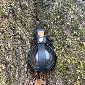 Puede incluir: Una peque&ntilde;a botella de vidrio transparente con un tap&oacute;n de corcho est&aacute; asegurada con una correa de cuero negro. La botella est&aacute; enclavada en un tronco de &aacute;rbol.