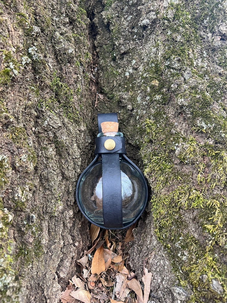 Puede incluir: Una peque&ntilde;a botella de vidrio transparente con un tap&oacute;n de corcho est&aacute; asegurada con una correa de cuero negro. La botella est&aacute; enclavada en un tronco de &aacute;rbol.