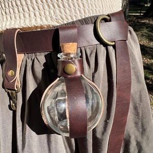 May include: A brown leather belt with a round metal buckle and a glass bottle attached to it. The bottle is held in place by a leather strap and has a cork stopper.