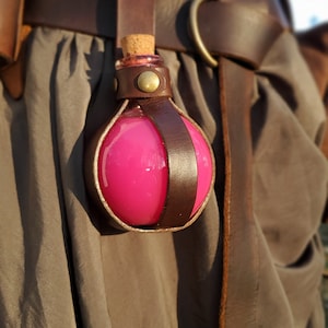 Puede incluir: Una botella de poci&oacute;n rosa con un tap&oacute;n de corcho, asegurada en un soporte de cuero marr&oacute;n. El soporte est&aacute; unido a un cintur&oacute;n de cuero marr&oacute;n.