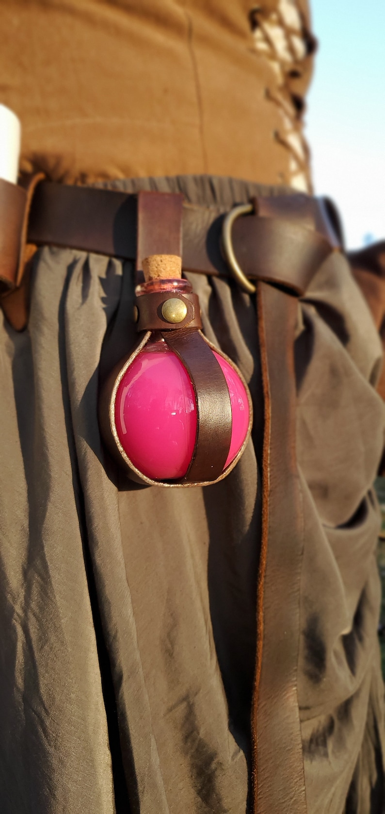 Puede incluir: Una botella de poci&oacute;n rosa con un tap&oacute;n de corcho, asegurada en un soporte de cuero marr&oacute;n. El soporte est&aacute; unido a un cintur&oacute;n de cuero marr&oacute;n.