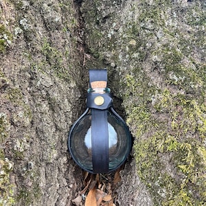 Puede incluir: Una peque&ntilde;a botella de vidrio transparente con un tap&oacute;n de corcho est&aacute; asegurada en un soporte de cuero negro. La botella est&aacute; enclavada en una grieta de un tronco de &aacute;rbol cubierto de musgo.