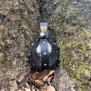 Puede incluir: Una botella de vidrio envuelta en cuero negro con un tap&oacute;n de corcho y detalles met&aacute;licos. La botella est&aacute; en un tronco de &aacute;rbol.