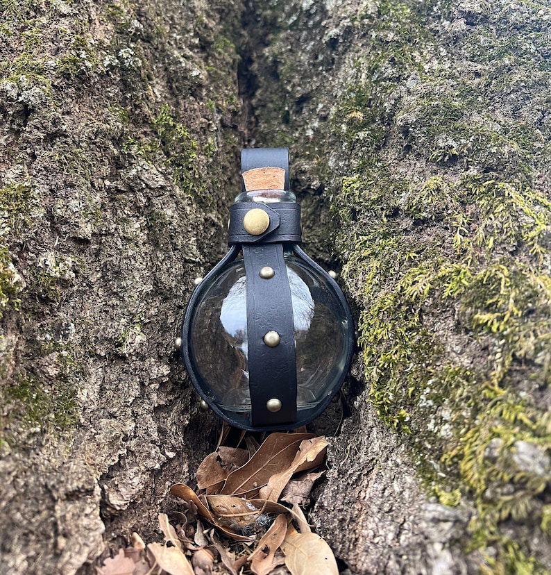 Puede incluir: Una botella de vidrio envuelta en cuero negro con un tap&oacute;n de corcho y detalles met&aacute;licos. La botella est&aacute; en un tronco de &aacute;rbol.
