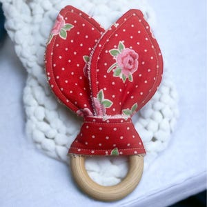 May include: A red fabric baby teether with a floral design and white polka dots. The teether has a wooden ring and a flower-shaped top. The fabric is red with white polka dots and pink roses. The teether is designed for babies.
