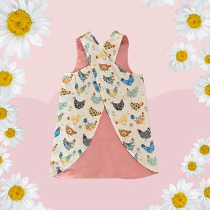 May include: A reversible apron with a cream chicken print and a solid pink reverse. The apron has a criss-cross back design and is decorated with colourful chickens and floral accents.