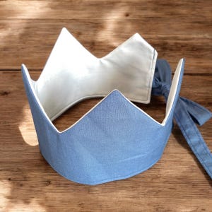 May include: A light blue fabric crown with a white interior and a matching blue tie. The crown has a simple, geometric design with three upward-pointing peaks. The crown is likely for a child or a pet.