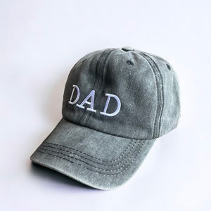 May include: A gray, distressed, cotton baseball cap with white embroidered text that reads "DAD".