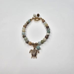 Peut inclure: Bracelet de perles avec un charm en forme de tortue de couleur dorée. Le bracelet est composé de diverses perles aux tons terreux, notamment beige, marron et bleu clair. Le bracelet a un fermoir doré. Un bijou charmant.