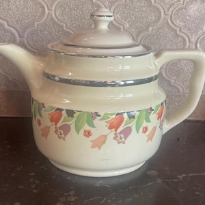 May include: A vintage cream-colored teapot with a silver trim and floral design. The teapot has a floral pattern with orange, pink, and purple flowers.