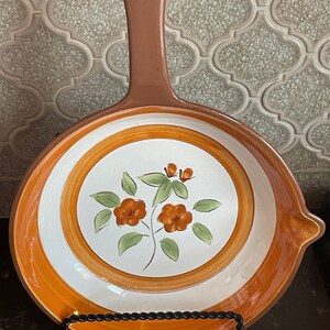 May include: A vintage orange and white ceramic frying pan with a floral design. The pan has a brown handle and a white rim.