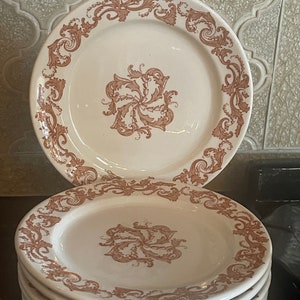 May include: A stack of white ceramic plates with a brown floral design. The plates have a decorative border and a central floral motif. The plates are stacked on a dark surface.