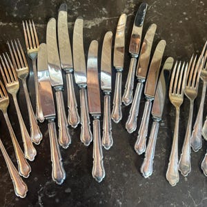 May include: An assortment of vintage silverware, including knives and forks, arranged on a dark surface. The knives have a shiny, silver blade and a decorative handle. The forks have four tines and a matching handle. The silverware appears to be antique.