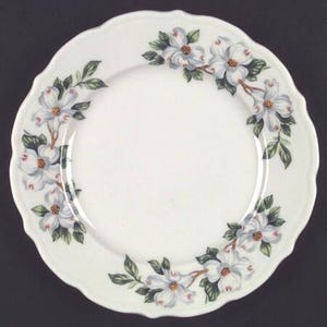 May include: White ceramic plate with a scalloped edge, adorned with a floral pattern of white dogwood blossoms and green leaves. The flowers are arranged around the rim of the plate, creating a decorative border.