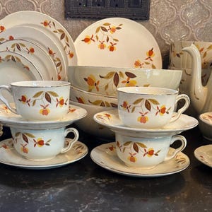 May include: A vintage cream-colored dinnerware set with a floral pattern in shades of orange, brown, and yellow. The set includes plates, bowls, teacups, saucers, and a teapot. The rims of the dishes have a gold trim.