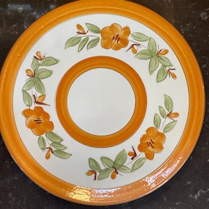 May include: A white ceramic plate with an orange rim and a floral design. The flowers are orange and yellow with green leaves. The plate has a center circle.