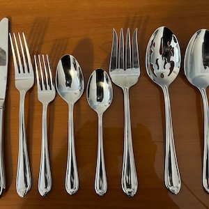 May include: A collection of polished silverware, including a knife, forks, spoons, and a serving spoon with a decorative cutout. The silverware has a reflective surface and a detailed handle design. The arrangement is set against a warm, wooden surface.
