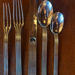 May include: A five-piece stainless steel flatware set featuring a small fork, a large fork, a dinner knife, a large spoon, and a small spoon. Each piece has a polished silver finish and a distinct handle design with three parallel vertical lines.