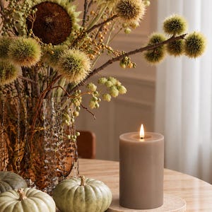 May include: A lit taupe pillar candle sits on a circular stone coaster next to two pale green pumpkins. Behind them, a textured amber glass vase holds a large arrangement of dried sunflowers, spiky chestnut husks, and green berries.