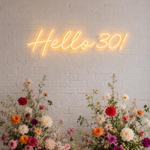 May include: A yellow neon sign displaying the text Hello 30! mounted on a white brick wall above two large floral arrangements featuring roses, dahlias, and various wildflowers in shades of pink, red, orange, white, and purple.