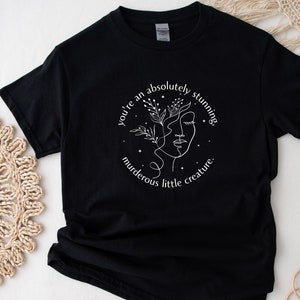 Op de afbeelding: Zwart T-shirt met een wit grafisch ontwerp van een gestileerd gezicht met bloemen en de tekst "You're an absolutely stunning murderous little creature."